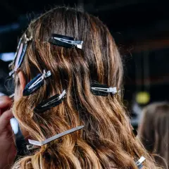 Detrás de cámaras, una maquilladora aplica producto para alisar el pelo de una modelo, sujetado con pinzas.