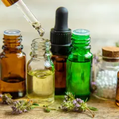 Gotas de aceite esencial cayendo de un gotero a una botella. Botellas de vidrio ámbar y verde, flores y un frasco con bolitas blancas. Ideal para aprender como hacer aceites esenciales caseros.