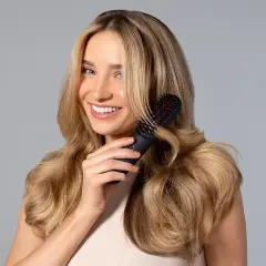 Mujer sonriente con cabello rubio ondulado usando un cepillo para rizar el pelo.