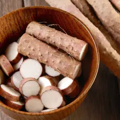 Un tazón de madera lleno de rodajas de ñame y dos tubérculos enteros. Detrás, varios tubérculos más.