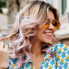 Mujer sonríe con cabello rubio ondulado y mechas rosas, luciendo gafas de sol naranjas. Un ejemplo de cómo reparar el pelo dañado con estilo.