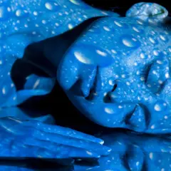 Una persona con la piel pintada de azul, cubierta de gotas blancas que parecen reflejar **que es la luz azul**.