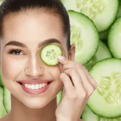 Una mujer sonríe mientras se coloca una rodaja de pepino en el ojo, rodeada de más rodajas de pepino.