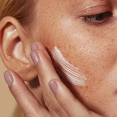 Una mano aplica crema en la mejilla de una mujer con pecas. La crema, que ayuda a controlar el sebo, deja un rastro blanco.