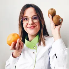 Mujer con gafas sostiene una naranja y dos kiwis, mostrando alimentos buenos para la piel.