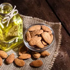 Botella de aceite de almendras para el pelo junto a un cuenco de almendras enteras sobre tela de arpillera.