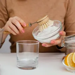 Manos con manicura rosa preparan una mezcla de agua con bicarbonato para limpiar. Se ve un vaso, limón y un cepillo.