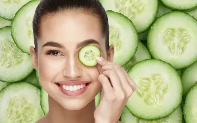 Una mujer sonríe mientras se coloca una rodaja de pepino en el ojo, rodeada de más rodajas de pepino.