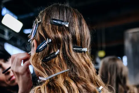 Detrás de cámaras, una maquilladora aplica producto para alisar el pelo de una modelo, sujetado con pinzas.