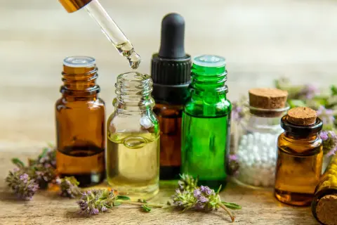 Gotas de aceite esencial cayendo de un gotero a una botella. Botellas de vidrio ámbar y verde, flores y un frasco con bolitas blancas. Ideal para aprender como hacer aceites esenciales caseros.
