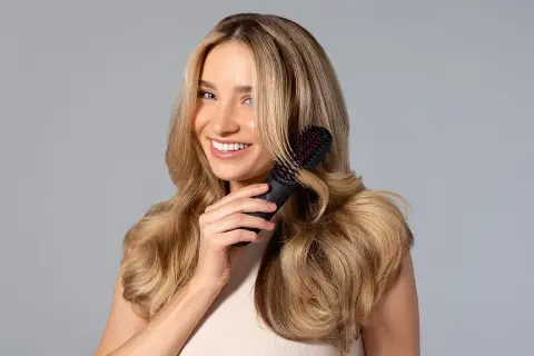 Mujer sonriente con cabello rubio ondulado usando un cepillo para rizar el pelo.