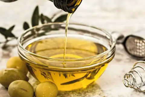 Aceite vegetal dorado se vierte en un cuenco, junto a aceitunas y utensilios de cocina.