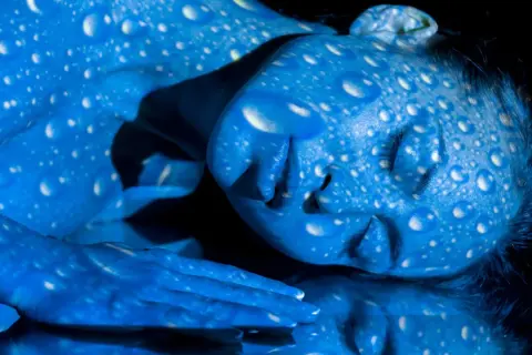 Una persona con la piel pintada de azul, cubierta de gotas blancas que parecen reflejar **que es la luz azul**.