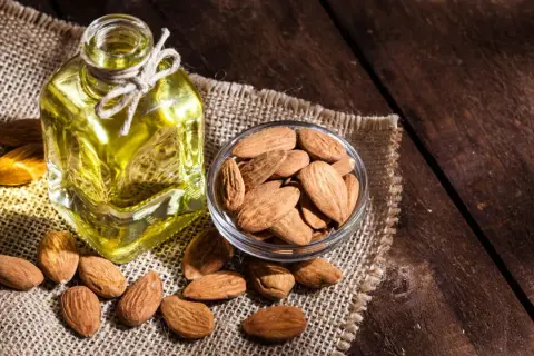 Botella de aceite de almendras para el pelo junto a un cuenco de almendras enteras sobre tela de arpillera.