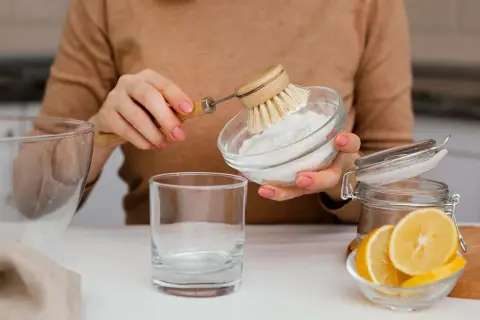 Manos con manicura rosa preparan una mezcla de agua con bicarbonato para limpiar. Se ve un vaso, limón y un cepillo.