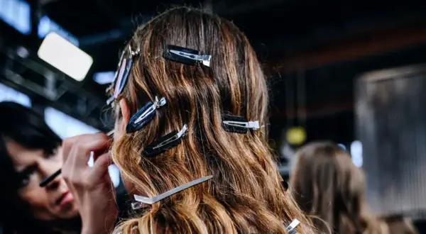 Detrás de cámaras, una maquilladora aplica producto para alisar el pelo de una modelo, sujetado con pinzas.