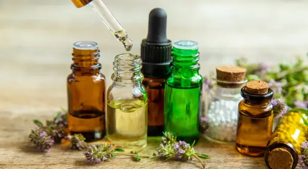 Gotas de aceite esencial cayendo de un gotero a una botella. Botellas de vidrio ámbar y verde, flores y un frasco con bolitas blancas. Ideal para aprender como hacer aceites esenciales caseros.