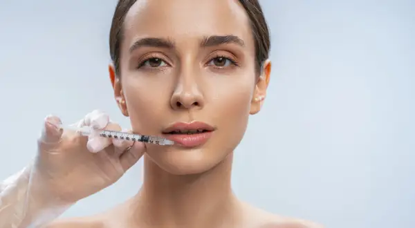 Una mano enguantada aplica ácido hialurónico en los labios de una mujer para darles volumen y definición.