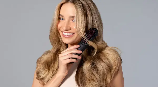 Mujer sonriente con cabello rubio ondulado usando un cepillo para rizar el pelo.