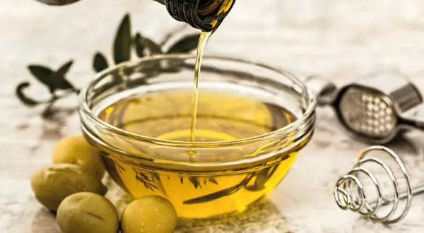 Aceite vegetal dorado se vierte en un cuenco, junto a aceitunas y utensilios de cocina.