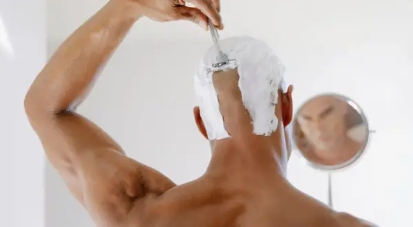 Persona se afeita la cabeza con espuma y cuchilla, reflejada en un espejo. Un ejemplo de como raparse la cabeza mujer.