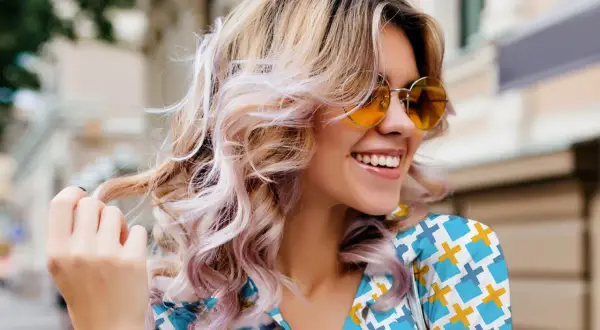 Mujer sonríe con cabello rubio ondulado y mechas rosas, luciendo gafas de sol naranjas. Un ejemplo de cómo reparar el pelo dañado con estilo.
