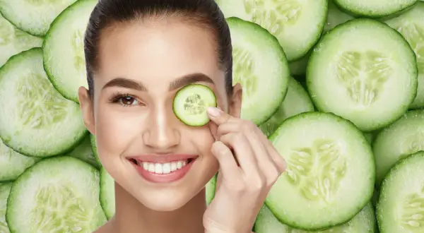 Una mujer sonríe mientras se coloca una rodaja de pepino en el ojo, rodeada de más rodajas de pepino.