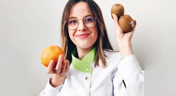 Mujer con gafas sostiene una naranja y dos kiwis, mostrando alimentos buenos para la piel.
