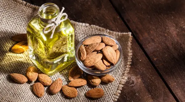 Botella de aceite de almendras para el pelo junto a un cuenco de almendras enteras sobre tela de arpillera.