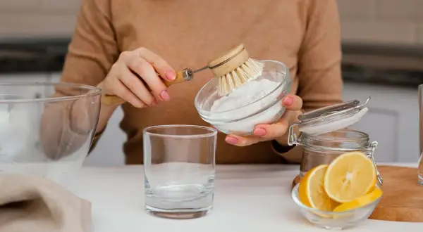 Manos con manicura rosa preparan una mezcla de agua con bicarbonato para limpiar. Se ve un vaso, limón y un cepillo.