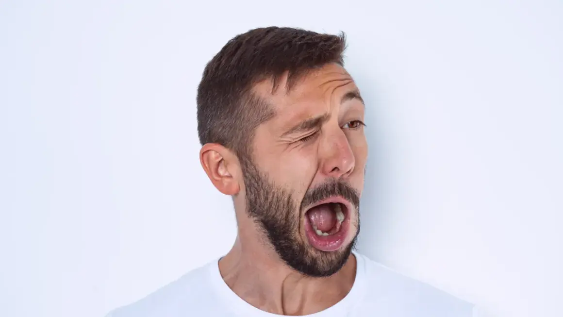Hombre con barba y tics nerviosos musculares, guiñando un ojo y con la boca abierta en un gesto de sorpresa o grito.