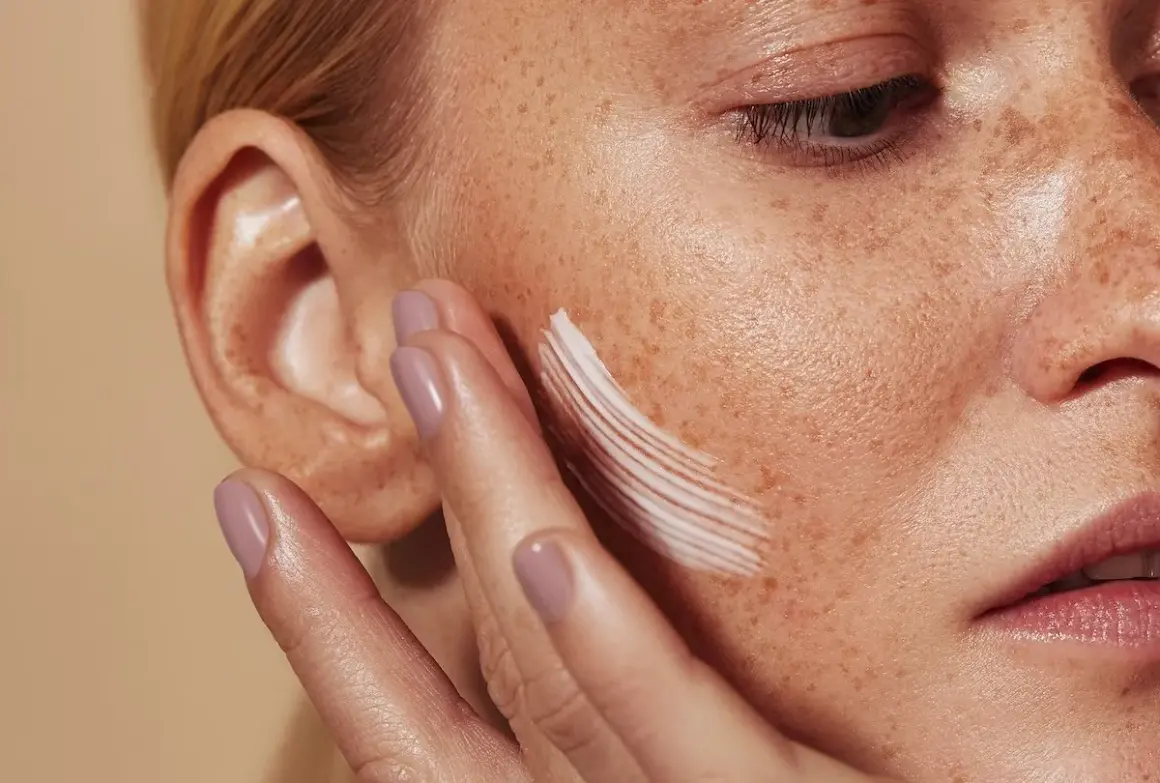 Una mano aplica crema en la mejilla de una mujer con pecas. La crema, que ayuda a controlar el sebo, deja un rastro blanco.