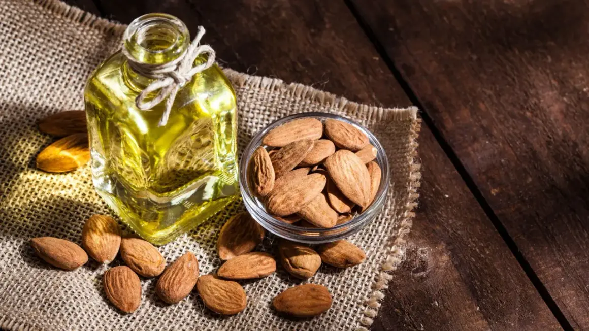 Botella de aceite de almendras para el pelo junto a un cuenco de almendras enteras sobre tela de arpillera.