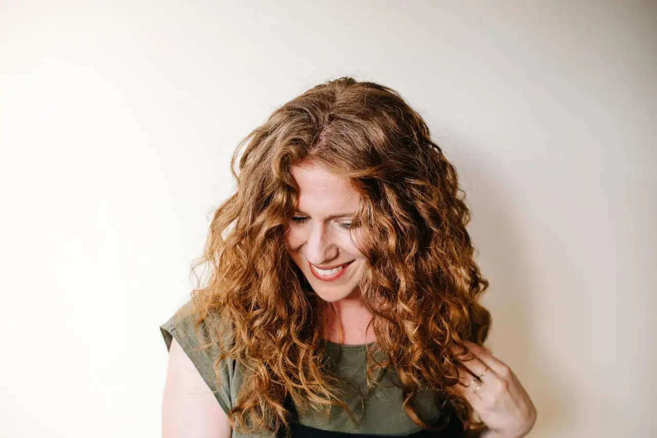 Mujer con cabello pelirrojo y rizado, sonriendo y mirando hacia abajo. Un ejemplo de c&oacute;mo rizar el pelo de forma natural.