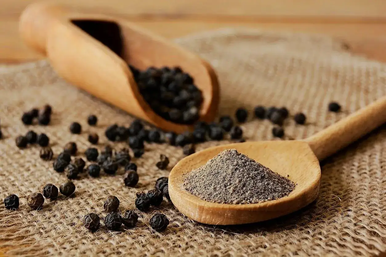 Cuchara de madera con pimienta negra molida, rodeada de granos enteros. Descubre las pimienta negra propiedades y su uso en la cocina.