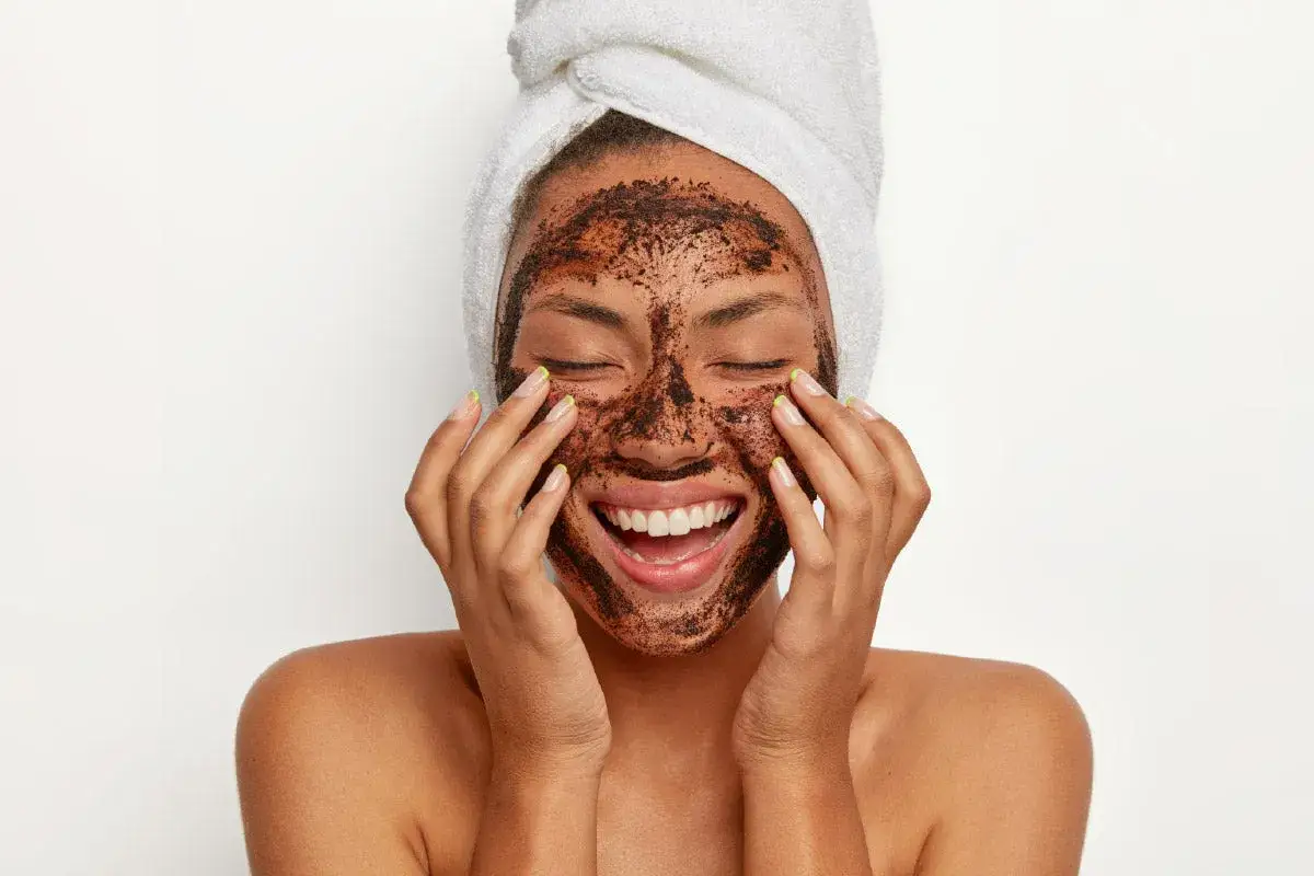 Mujer sonriente con mascarilla de café en el rostro, aprendiendo como exfoliar la piel para un brillo natural.