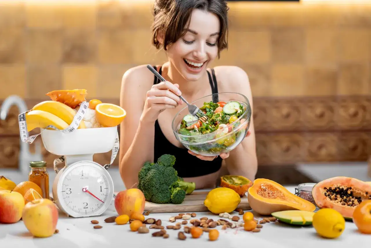 Mujer sonriente comiendo ensalada, rodeada de frutas y verduras. Una b&aacute;scula y cinta m&eacute;trica sugieren que est&aacute; pensando en qu&eacute; hacer para engordar de forma saludable.