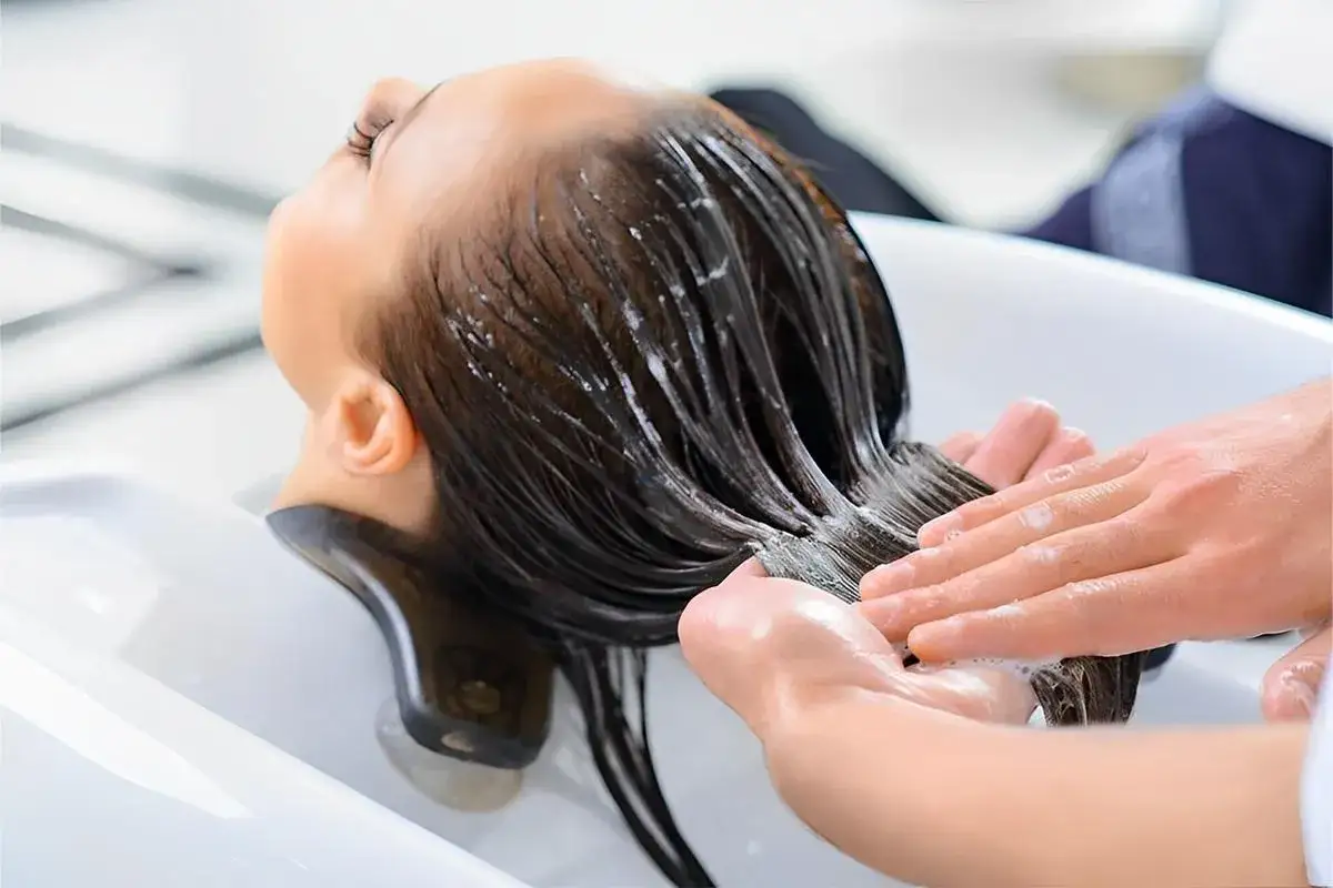 Manos aplican acondicionador en el cabello mojado de una mujer en un lavabo de peluquer&iacute;a. El acondicionador sirve para suavizar y nutrir el pelo.