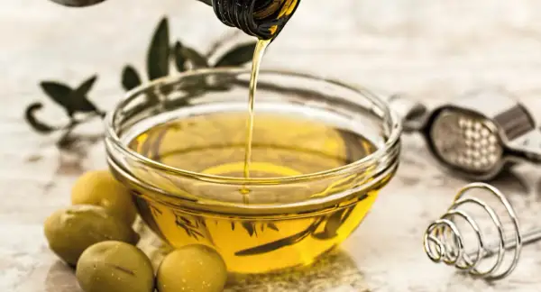 Aceite vegetal dorado se vierte en un cuenco, junto a aceitunas y utensilios de cocina.
