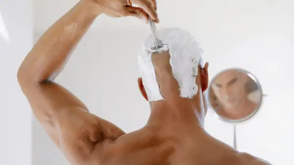 Persona se afeita la cabeza con espuma y cuchilla, reflejada en un espejo. Un ejemplo de como raparse la cabeza mujer.