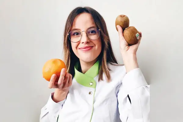 Mujer con gafas sostiene una naranja y dos kiwis, mostrando alimentos buenos para la piel.